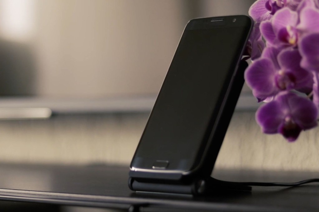 黒いスマートフォンがスタンドに立てかけられ、背景には紫の花が置かれています。シンプルなデザインで、モダンな雰囲気を醸し出しています。