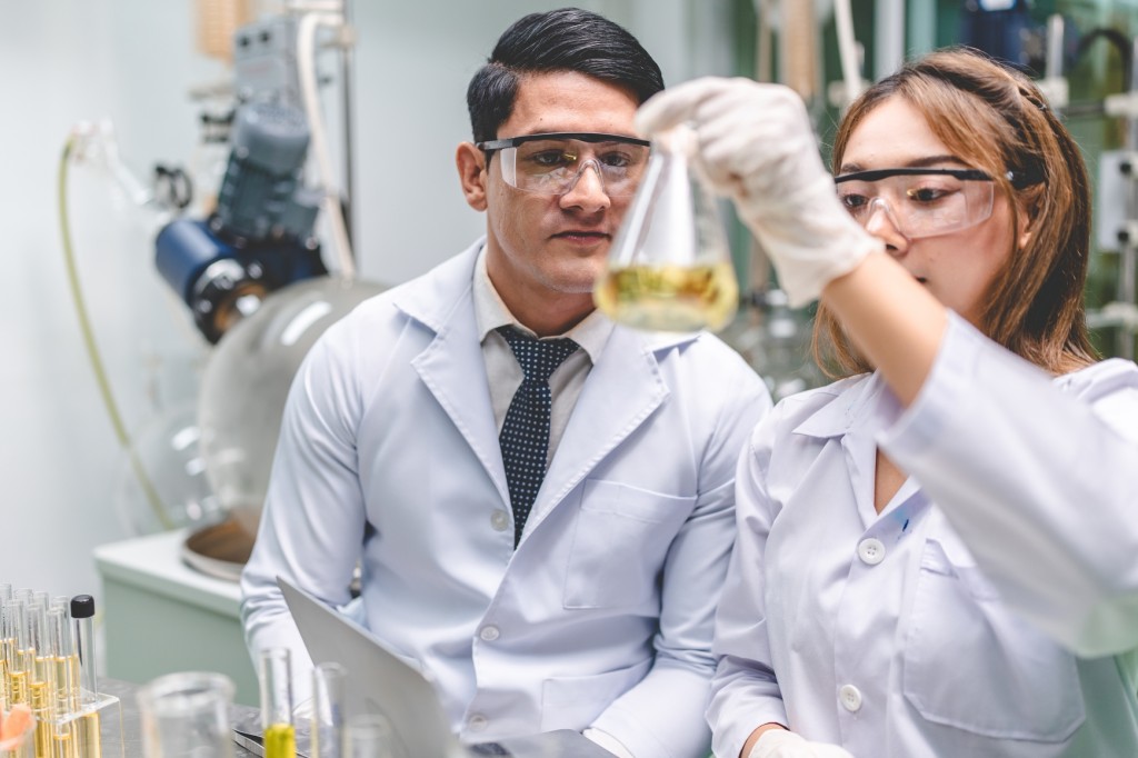 Two lab technicians in white lab coats and safety goggles looking at a yellow liquid in a glass flask