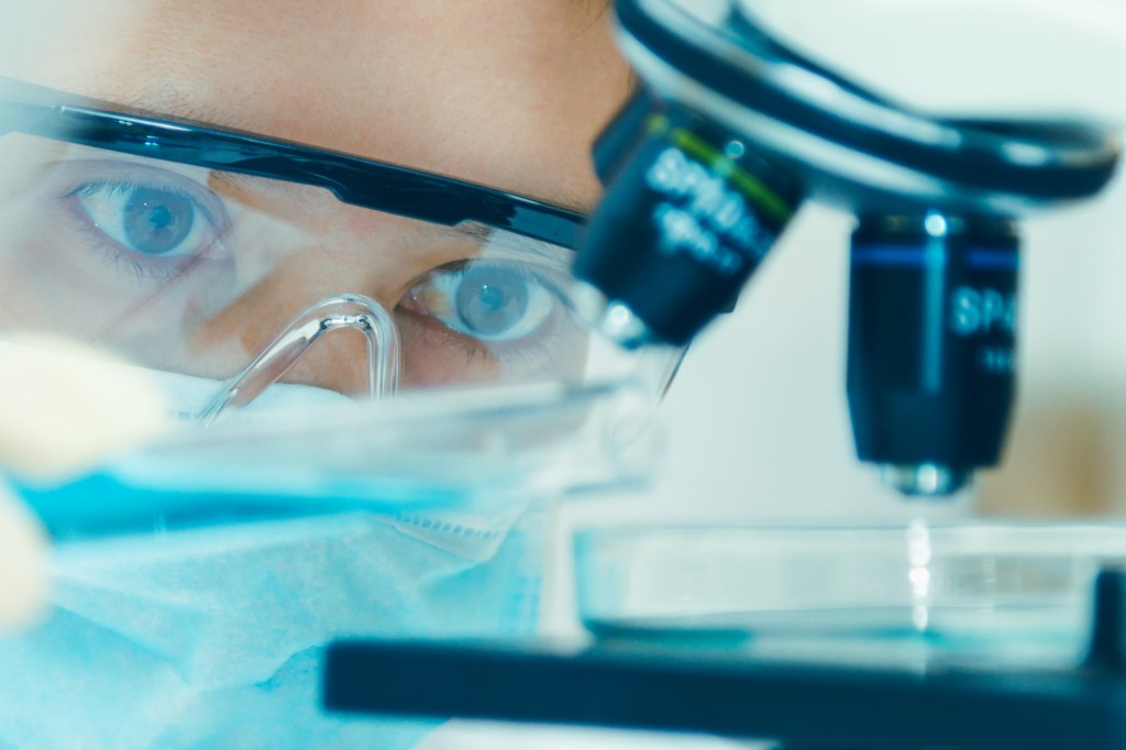 Close-up of person with safety goggles and mask looking at a petri dish under a microscope 