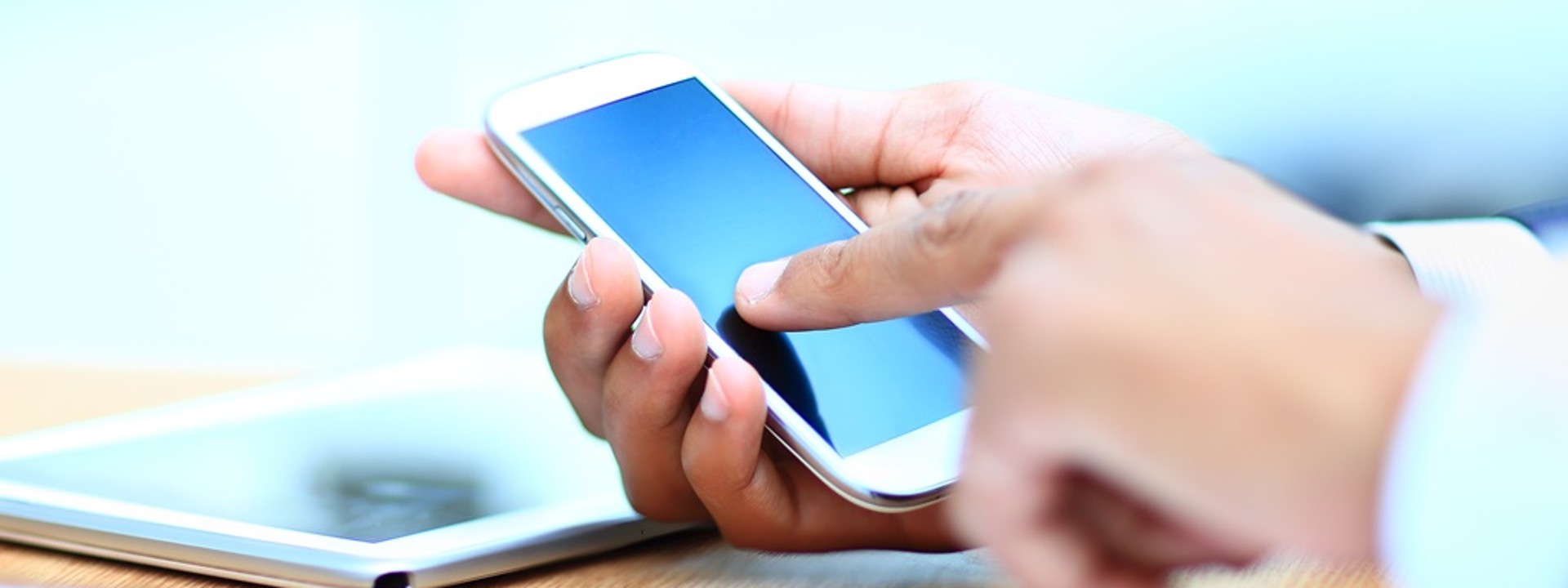 Close up of a person holding a white smartphone in their right hand and pointing to the phone screen with their left hand. The background is light and obscure.