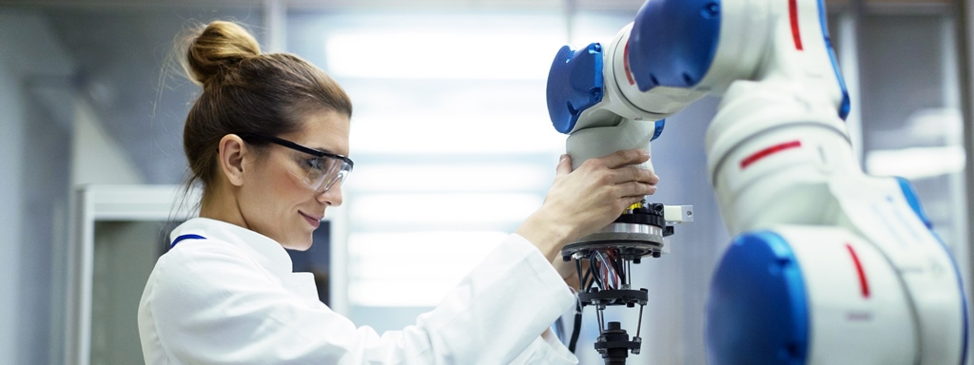 A feminine engineer adjusts a robotic arm in a laboratory which has wires hanging out of the end. She is wearing a white lab coat with safety glasses.