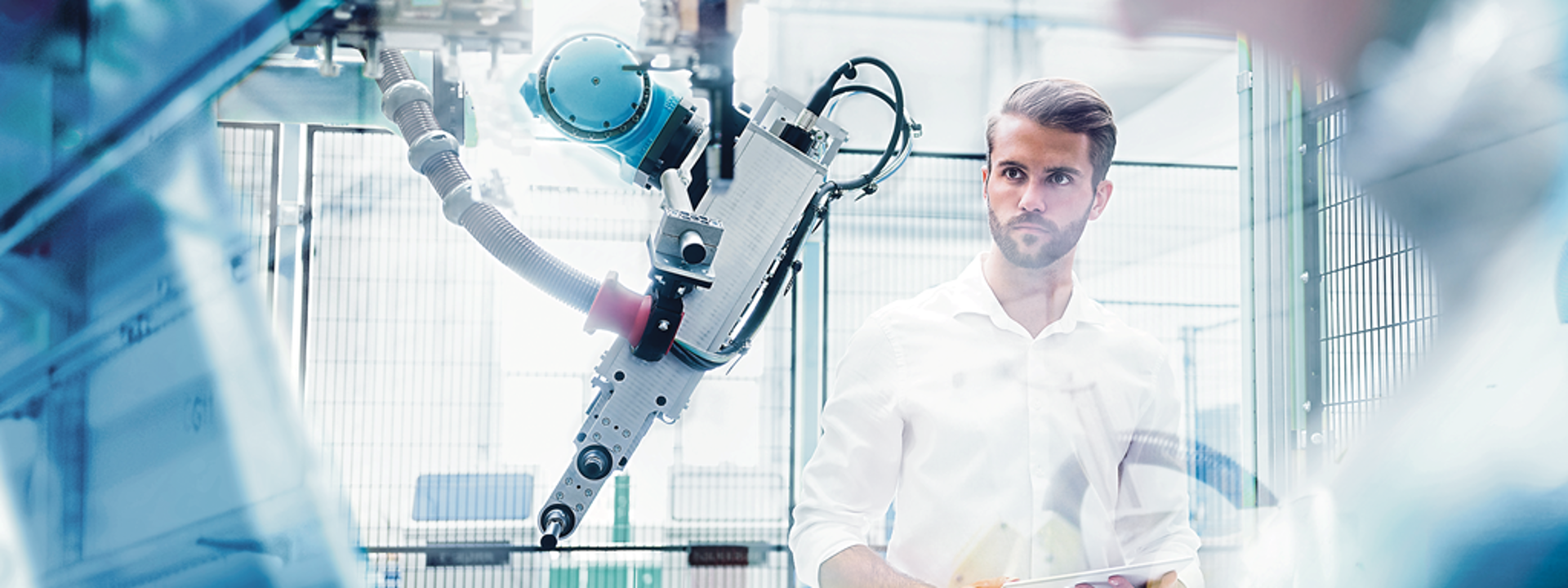 A casual engineer looking at a robotic arm from a safe distance holding a tablet.
