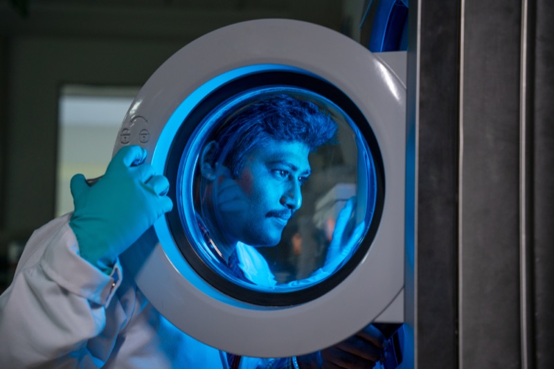 Labortechniker blickt durch das runde Fenster einer blau beleuchteten Prüfmaschine