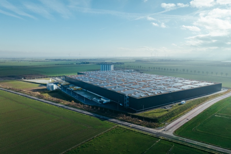 Luftaufnahme eines großen Industriegebäudes mit Solarpanels auf dem Dach umgeben von grünen Feldern