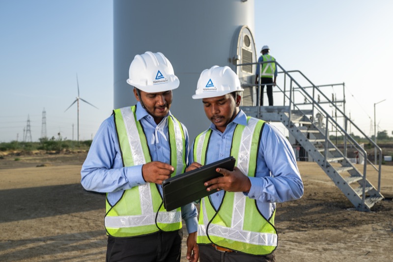 Two engineers in safety vests and hard hats reviewing a tablet near a wind turbine