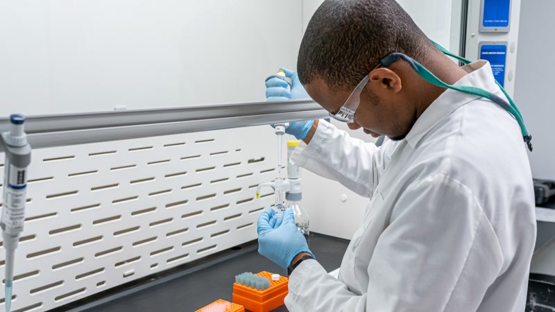 Laboratory technician in a white coat and safety goggles handling samples under a fume hood