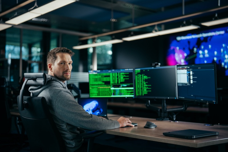 Man sitting at a desk with multiple monitors displaying lines of code in a dark office